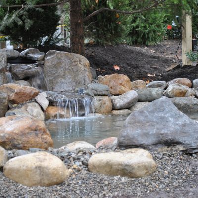A small artificial waterfall flows over rocks into a shallow pond, surrounded by stones and trees in a landscaped garden.