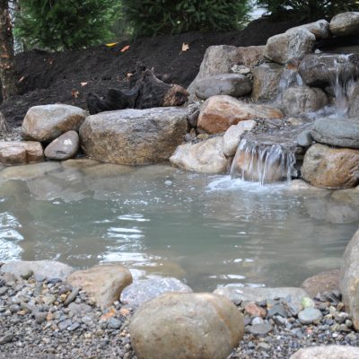 Small backyard pond with rocks and a gentle waterfall, surrounded by soil, pebbles, and evergreen trees in the background.