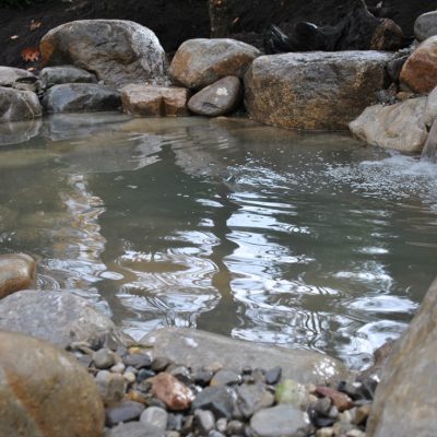 A small pond with clear water, surrounded by large rocks and two small waterfalls flowing into it.