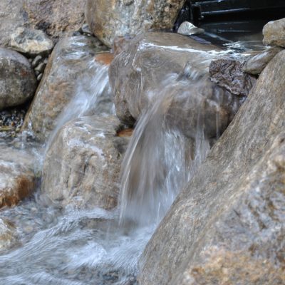 Clear water flows over large brown rocks in a small stream, creating a gentle waterfall among the stones.