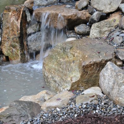 Small waterfall flows over rocks into a shallow, rocky pond; water meter visible at the upper left corner.