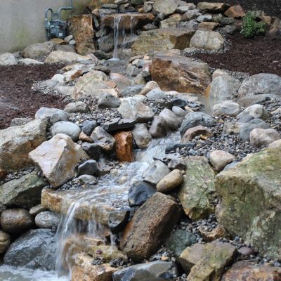 Water flows over rocks in a backyard landscape, creating a small artificial waterfall and stream.