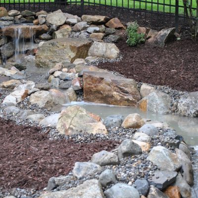 A small artificial stream with rocks and mulch flows through a landscaped backyard area near a fence.