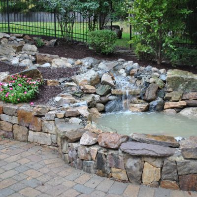 Small backyard pond with waterfall feature, surrounded by rocks, mulch, flowers, and stone paver patio.