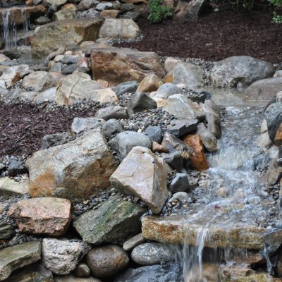Small artificial stream flows over rocks in a landscaped garden with mulch and various sized stones.