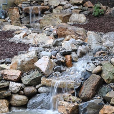 Small waterfall cascading over rocks in a landscaped garden with mulch and plants in the background.