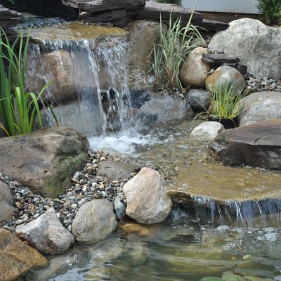 Small waterfall flows over rocks into a pond, surrounded by plants and smooth stones in a landscaped garden.