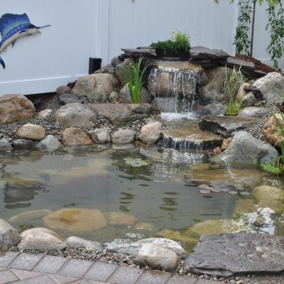 Small backyard pond with rocks, plants, and a waterfall feature; a decorative fish is mounted on the white fence.
