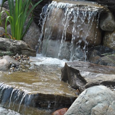 Small waterfall flows over rocks into a pond, surrounded by stones and green plants in a garden setting.