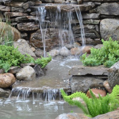 Small waterfall flows over rocks into a pond, surrounded by green plants and landscaping stones in a garden setting.