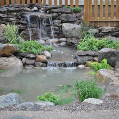 Small backyard pond with rocks, plants, and a cascading waterfall, surrounded by a wooden fence and gravel pathway.