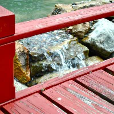 Red wooden railing and deck overlooking rocks with a small stream of water flowing between them.