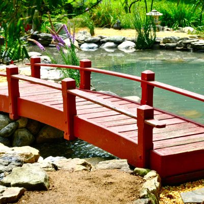 A small red wooden bridge crosses over a pond surrounded by rocks and greenery in a garden.