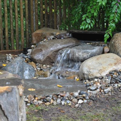 Small waterfall flows over rocks in a landscaped garden with a wooden fence and leafy green plants in the background.
