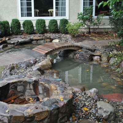 A small pond with a wooden footbridge and a stone fire pit in a landscaped backyard near a house with large windows.