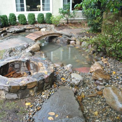A backyard with a stone fire pit, small pond, stream, and landscaping near a house with large windows.