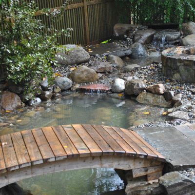 A small wooden bridge crosses over a pond with rocks and a waterfall in a fenced backyard garden.