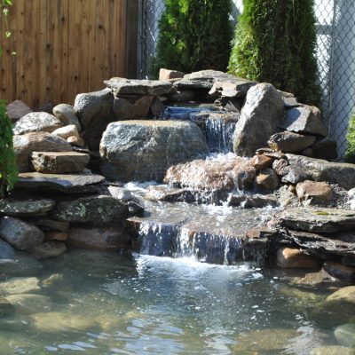 Small backyard waterfall flows over rocks into a pond, surrounded by trees and a wooden fence.