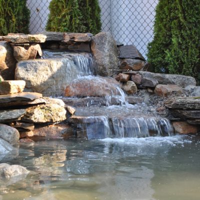 Small backyard waterfall with rocks and plants, flowing into a pond beside a chain-link fence and tall shrubs.
