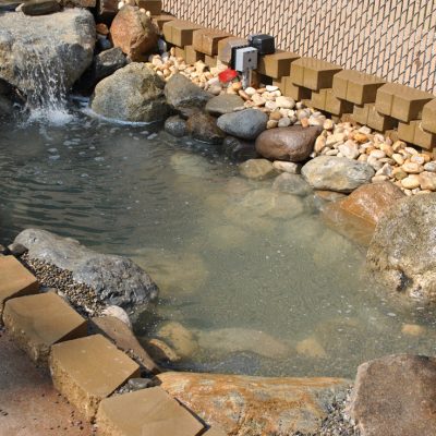 Small backyard pond with rocks, a waterfall feature, and a stone border next to a fence and gravel path.