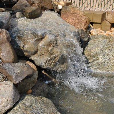 Water flows over rocks creating a small waterfall feature in a landscaped outdoor garden setting.