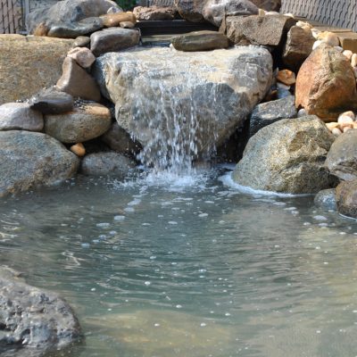 Small pond with clear water and a stone waterfall surrounded by large rocks in an outdoor setting.
