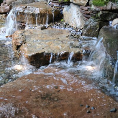 Water flows over flat rocks and small waterfalls in a landscaped garden stream with stones and moss in the background.