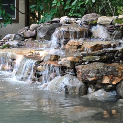 Small artificial waterfall flows over rocks into a clear pond, surrounded by greenery and a house in the background.