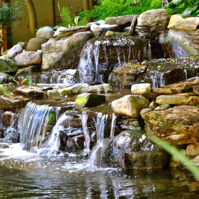 Small artificial waterfall flows over rocks into a pond, surrounded by green plants and stones in a garden setting.