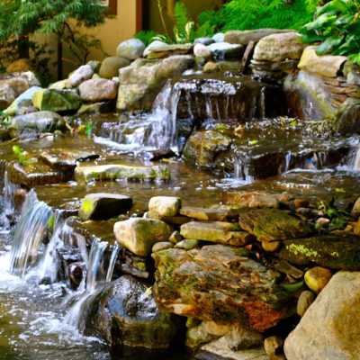 A small, multi-level stone waterfall flows into a pond, surrounded by green plants and rocks in a garden.
