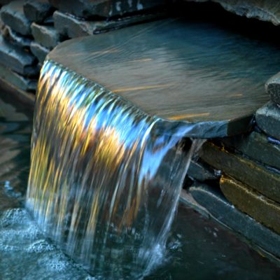Water cascades over a flat stone ledge into a small pool, with light reflecting colors on the surface of the waterfall.
