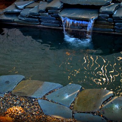 A small stone-lined pond with a short waterfall, surrounded by rocks and gravel, reflecting light on the water’s surface.