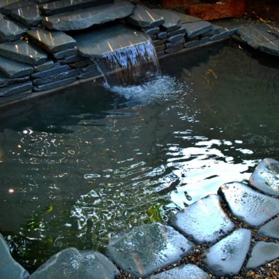Small backyard pond with stacked stone border and a short waterfall feature, surrounded by gravel.