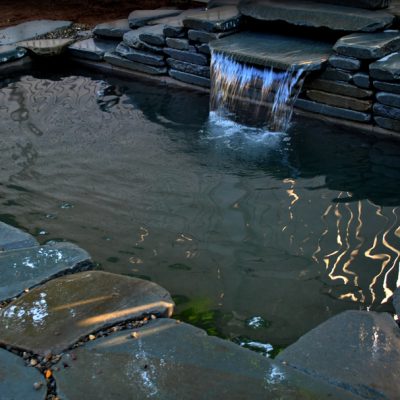 Small backyard pond with stone edging and a waterfall feature, reflecting light patterns on the water’s surface.
