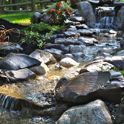 A small backyard stream flows over rocks near a flowerbed, with a hammock and wooden fence in the background.