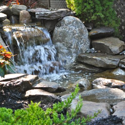 Small garden waterfall flows over rocks into a pond, surrounded by greenery and blooming plants.