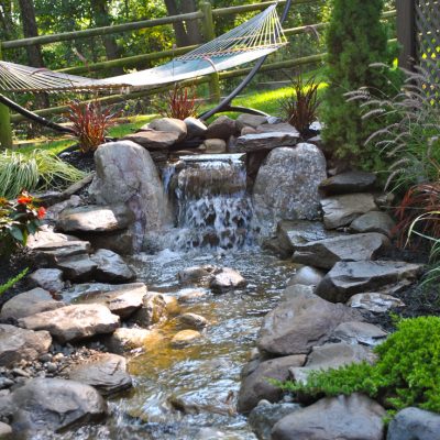 A small backyard waterfall flows over rocks, surrounded by plants and a hammock hanging in the background.