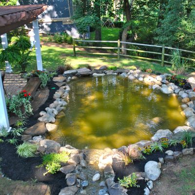 Backyard with a stone-edged pond, surrounding plants, patio seating, and wooden fence in a wooded area.