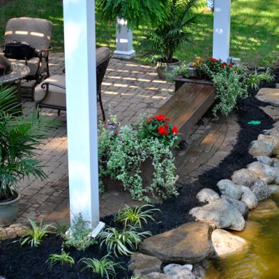 Patio with outdoor seating, potted plants, and a grill beside a rock-lined pond and garden.