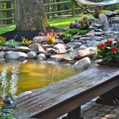 A garden pond with flowing water, surrounded by rocks, flowers, a wooden bench, and a hammock in the background.