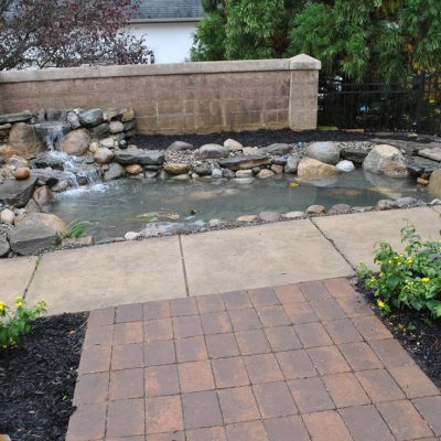 A small backyard pond with a stone waterfall feature, surrounded by a paved walkway and landscaped plants.