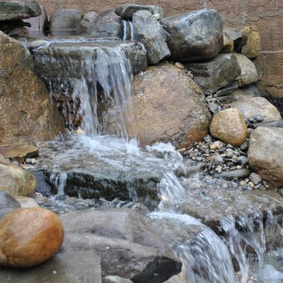 Water flows over rocks in a small backyard waterfall feature, with pebbles and large stones surrounding the stream.