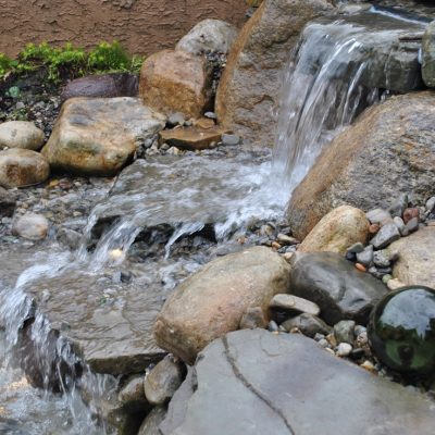 Water flows over rocks in a small landscaped backyard waterfall, surrounded by stones and pebbles.