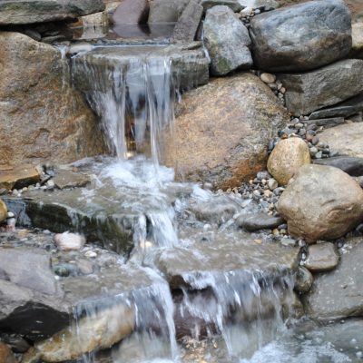 Water flows over a small stone waterfall surrounded by large rocks and pebbles in a natural setting.