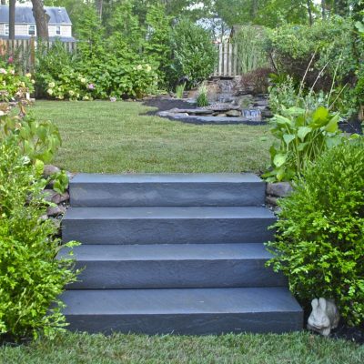 Gray stone steps lead up to a grassy yard, bordered by green bushes and garden plants on both sides.