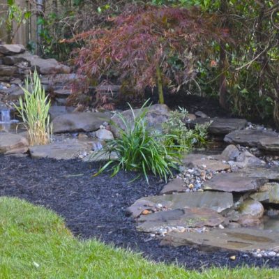 A backyard garden pond with small waterfalls, surrounded by rocks, plants, and fresh mulch along a grassy lawn.