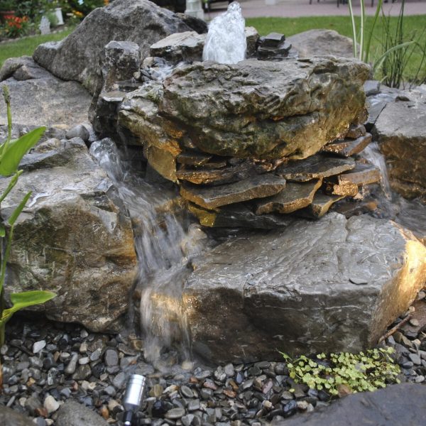 Small backyard water feature with cascading stream over stacked rocks, surrounded by plants and pebbles.