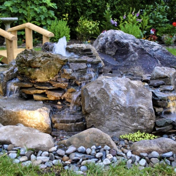 A small rock waterfall with a wooden footbridge, surrounded by stones, plants, and grass in a garden.