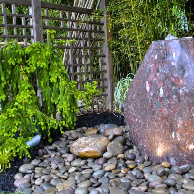 Large polished stone water feature surrounded by smooth rocks, greenery, and a wooden lattice fence in a garden setting.