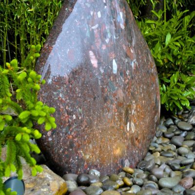 Large polished stone water fountain surrounded by smooth river rocks and green plants in a garden setting.